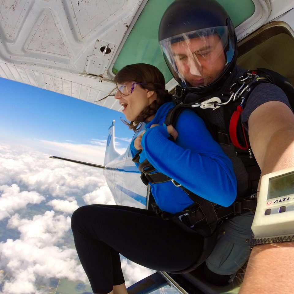 Female tandem skydiving student with blue shirt prepares to exit airplane with her instructor