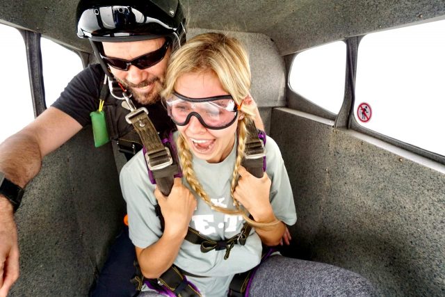 Blond tandem skydiving student with braids sits with instructor in plane on the way up to altitude to skydive