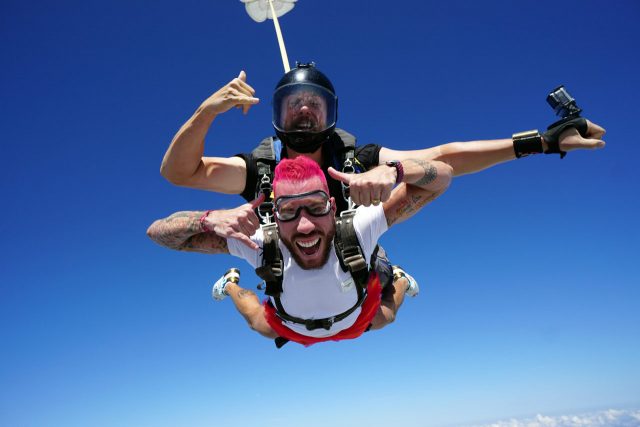 Male tandem skydiving student with pink hair smiles at camera while in freefall