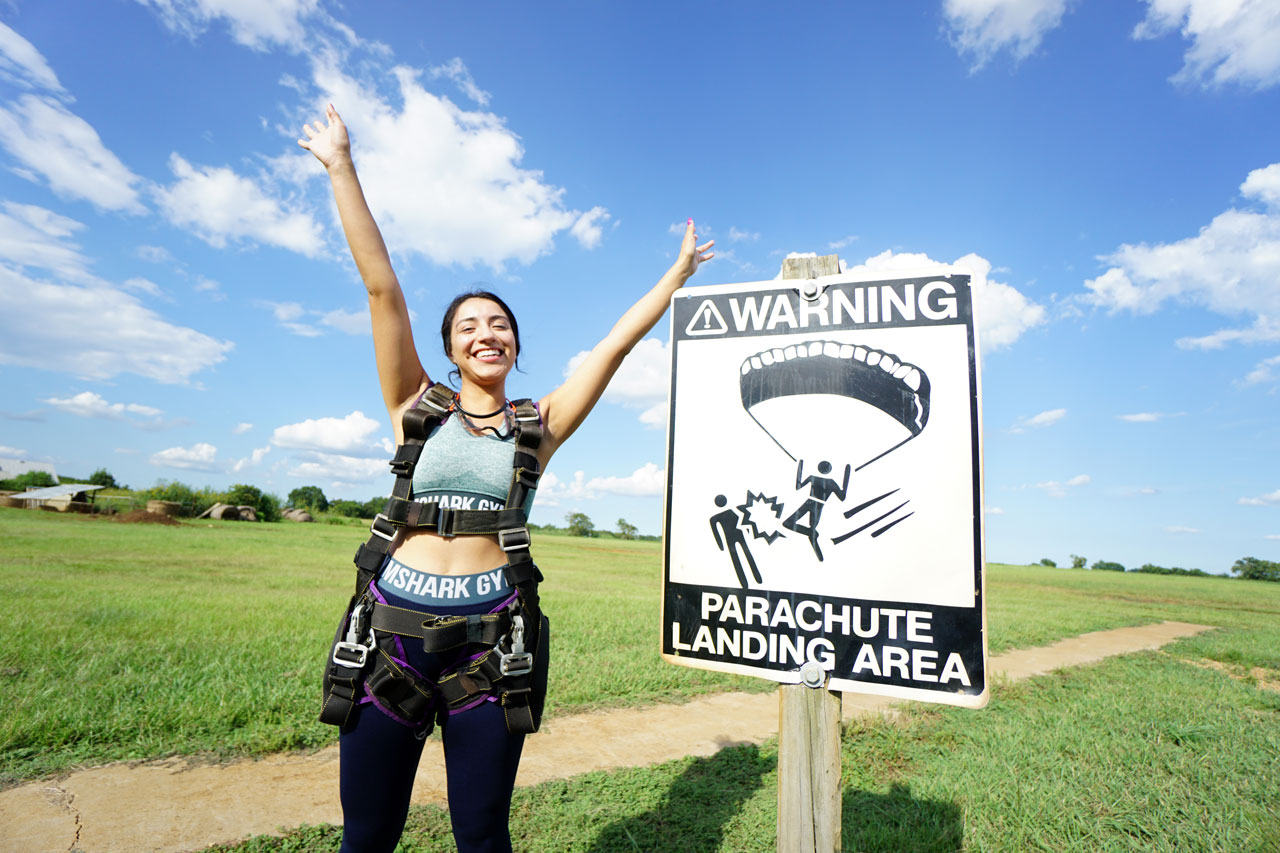 Young female tandem student with hands in the air poses next to a sign reading "parachute landing area" at Texas Skydiving