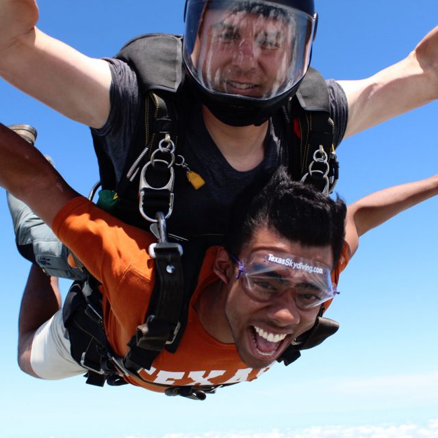 Young man wearing an orange t-shirt smiles at camera during a tandem skydive at Texas Skydiving near College Station, TX