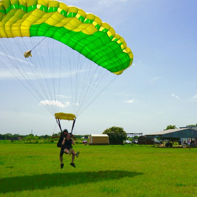 Tandem skydiving student and instructor under a green and yellow parachute landing in a green field at Texas Skydiving