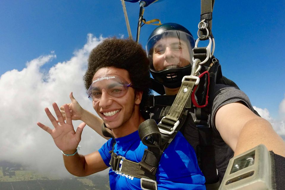 Smiling man with dark hair waves at camera while under canopy during a tandem skydive at Texas Skydiving near Austin, TX